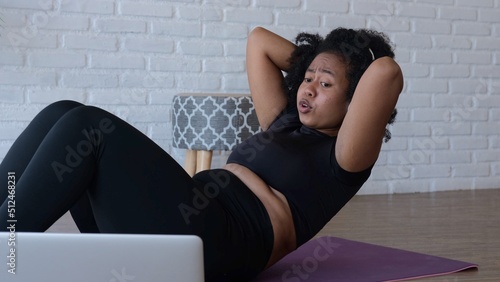 An Afro-Asiatic woman in a bright room lying on a sports mat diligently pumps the press with proper breathing and does not strain her neck for a beautiful and toned body. Fitness online. photo