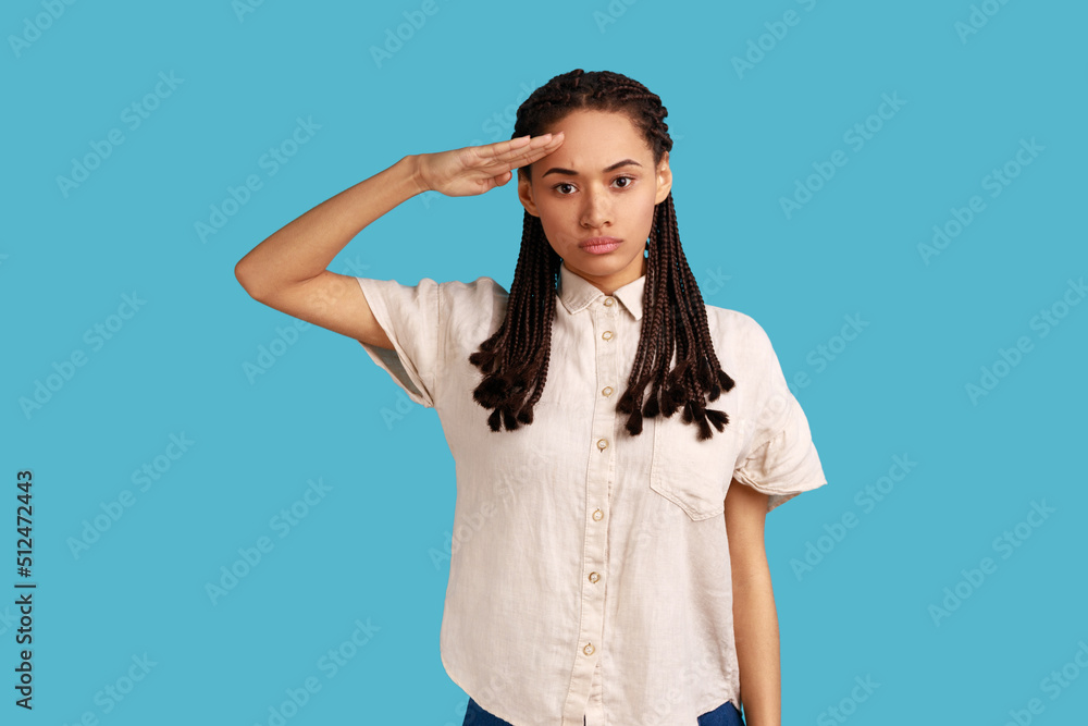 Yes sir. Portrait of responsible serious woman with black dreadlocks saluting commander, listening order with obedient expression, wearing white shirt. Indoor studio shot isolated on blue background.