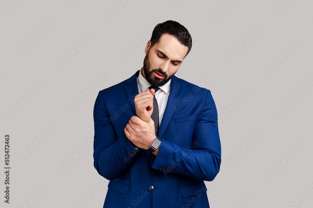 Portrait of businessman standing with grimace of pain, massaging sore wrist, suffering hand injury or sprain, wearing official style suit. Indoor studio shot isolated on gray background. Portrait of businessman standing with grimace of pain, massaging sore wrist, suffering hand injury or sprain, wearing official style suit. Indoor studio shot isolated on gray background.