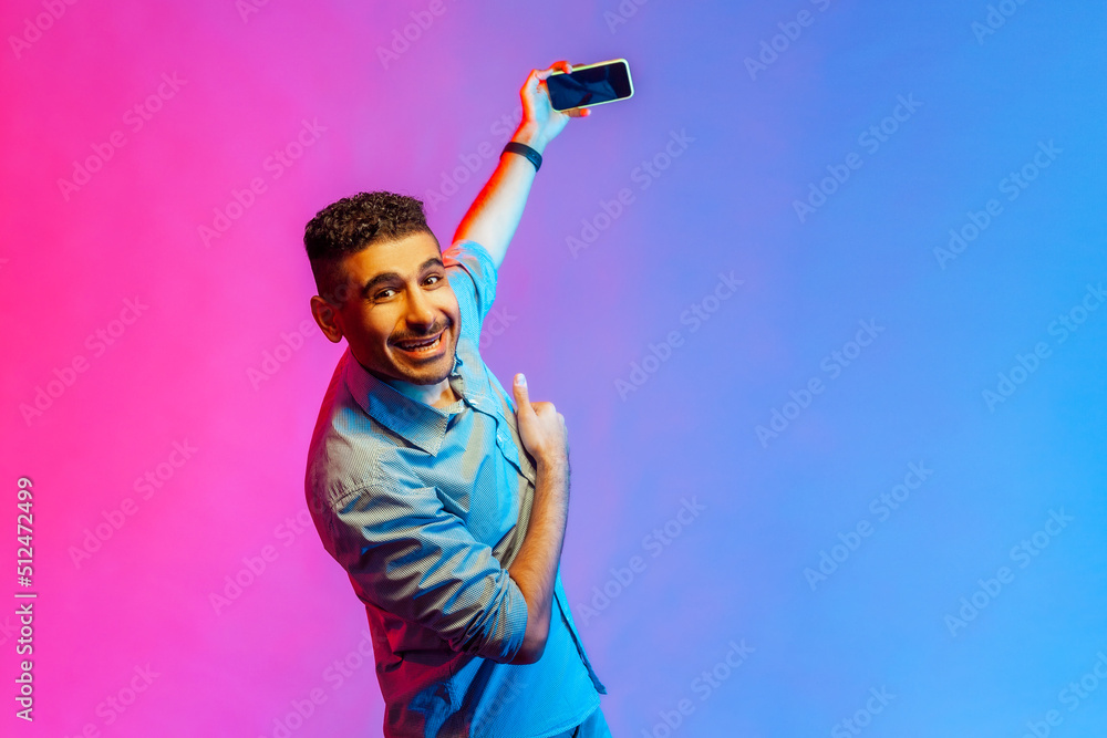 Portrait of smiling happy man in shirt standing with smart phone in hands, broadcasting livestream, looking at camera. Indoor studio shot isolated on colorful neon light background. Portrait of smiling happy man in shirt standing with smart phone in hands, broadcasting livestream, looking at camera. Indoor studio shot isolated on colorful neon light background.