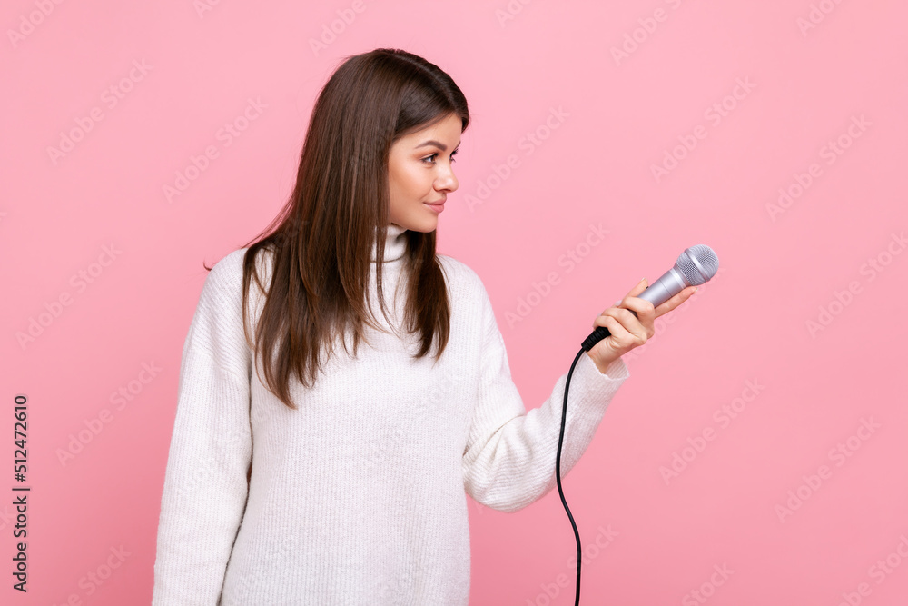 Side view portrait of brunette female offering microphone to somebody, journalist asking opinion, wearing white casual style sweater. Indoor studio shot isolated on pink background. Side view portrait of brunette female offering microphone to somebody, journalist asking opinion, wearing white casual style sweater. Indoor studio shot isolated on pink background.