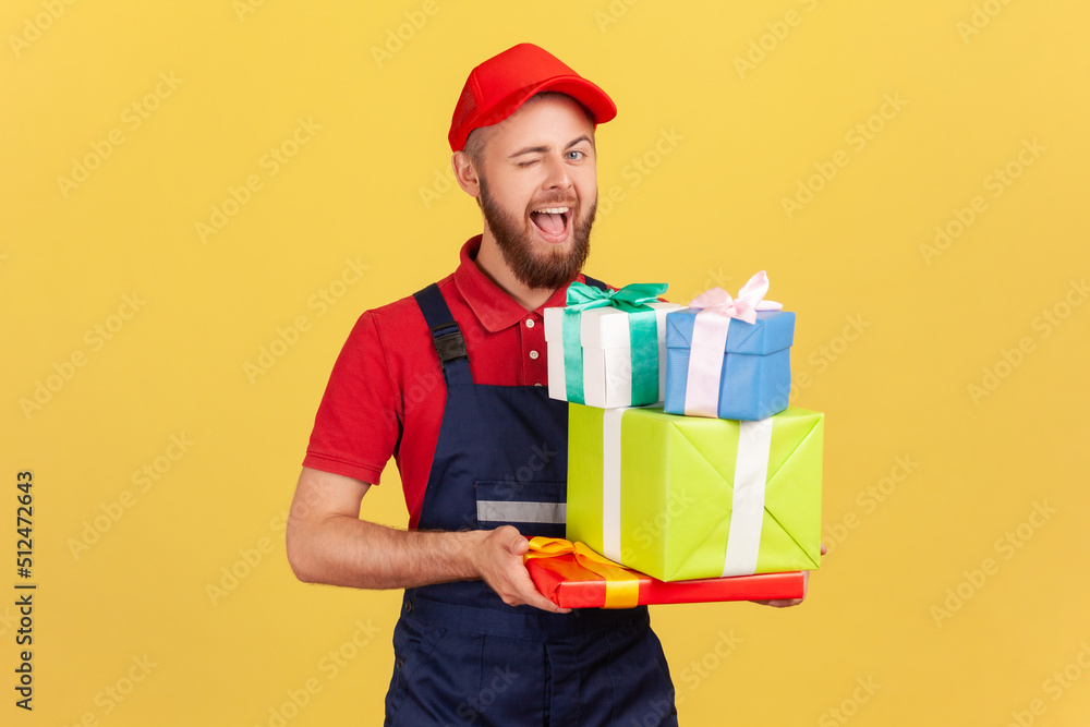 Portrait of playful childish courier man wearing uniform and cap holding many present boxes, bringing order for celebrating holiday, winking to camera. Indoor studio shot isolated on yellow background Portrait of playful childish courier man wearing uniform and cap holding many present boxes, bringing order for celebrating holiday, winking to camera. Indoor studio shot isolated on yellow background