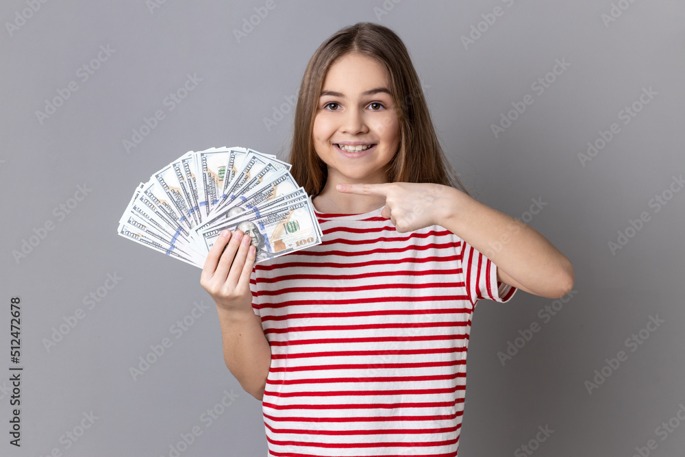Portrait of satisfied little girl wearing striped T-shirt pointing at fan of dollar banknotes, showing big money and looking at camera. Indoor studio shot isolated on gray background. Portrait of satisfied little girl wearing striped T-shirt pointing at fan of dollar banknotes, showing big money and looking at camera. Indoor studio shot isolated on gray background.