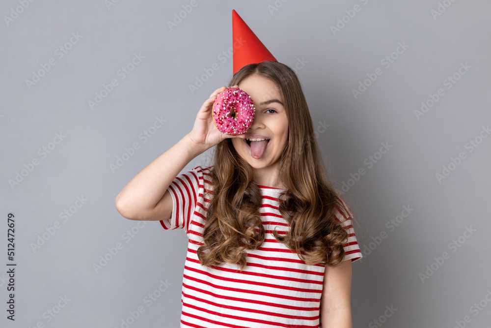 Portrait of playful joyful little girl wearing striped T-shirt and party cone, covering her eyes with tasty sweet donut, showing tongue out. Indoor studio shot isolated on gray background. Portrait of playful joyful little girl wearing striped T-shirt and party cone, covering her eyes with tasty sweet donut, showing tongue out. Indoor studio shot isolated on gray background.