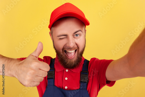 Satisfied smiling worker man wearing blue uniform taking selfie, looking at camera winking and showing thumb up, point of view of photo. Indoor studio shot isolated on yellow background.