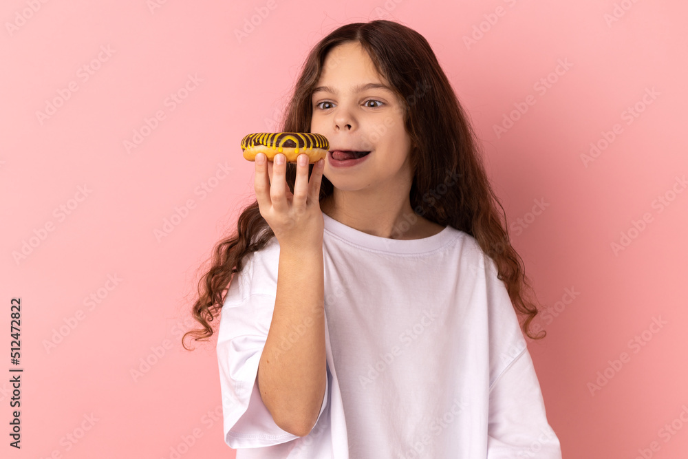 Portrait of delighted cute little girl wearing white T-shirt licking delicious donut, looking with desire to eat sweet dessert, showing tongue out. Indoor studio shot isolated on pink background. Portrait of delighted cute little girl wearing white T-shirt licking delicious donut, looking with desire to eat sweet dessert, showing tongue out. Indoor studio shot isolated on pink background.