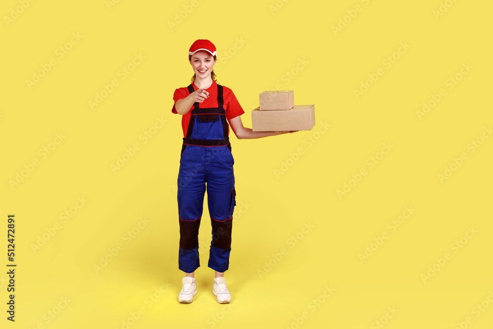 Full length portrait of positive courier woman pointing at camera, holding cardboard packages, fast delivery service, wears overalls and red cap. Indoor studio shot isolated on yellow background. Full length portrait of positive courier woman pointing at camera, holding cardboard packages, fast delivery service, wears overalls and red cap. Indoor studio shot isolated on yellow background.