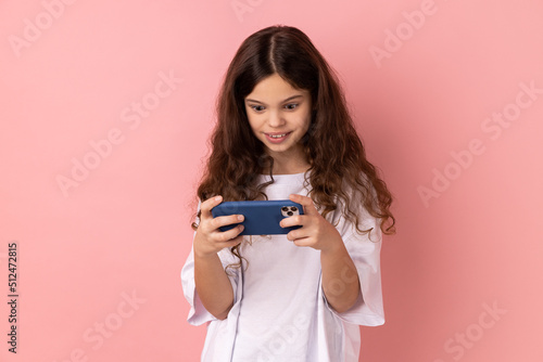 Portrait of surprised little girl wearing white T-shirt using mobile phone with shocked positive expression, playing video game on cellphone. Indoor studio shot isolated on pink background.