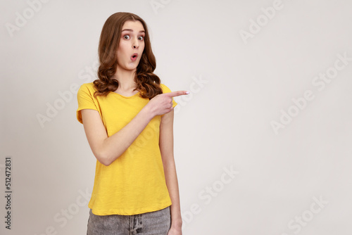 Portrait of amazed speechless pretty teenage girl collage student  standing with drop jaw and wondered  pointing at fascinating product. Indoor studio shot isolated on gray background.