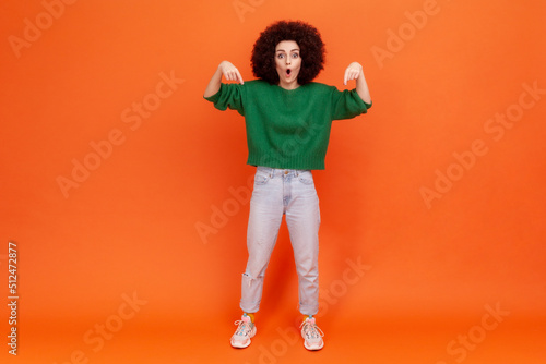 Full length portrait of excited smiling woman with Afro hairstyle in green casual sweater pointing fingers down, presenting space for advertisement. Indoor studio shot isolated on orange background.