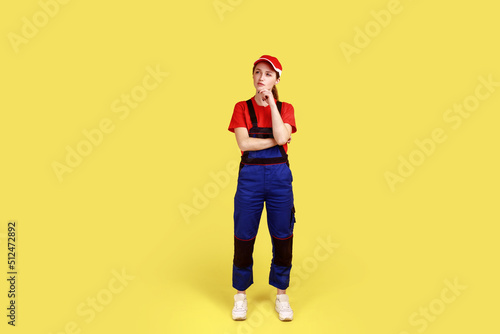 Full length portrait of pensive woman worker standing and holding chin, having thoughtful facial expression, wearing overalls and red cap. Indoor studio shot isolated on yellow background.