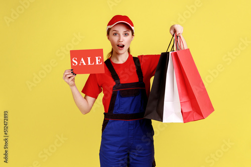 Portrait of surprised amazed delivery woman standing with shopping bags and card with sale inscription, wearing overalls and red cap. Indoor studio shot isolated on yellow background.