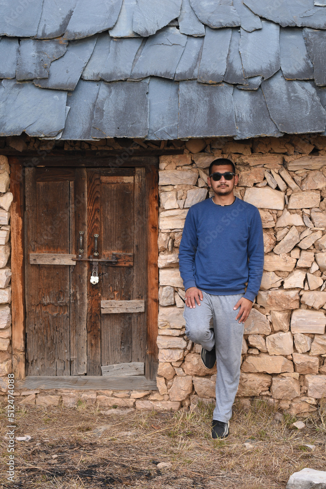 Man standing leaning against old structure wall made of stone and mud mixture looking straight wearing black sunglasses Man standing leaning against old structure wall made of stone and mud mixture looking straight wearing black sunglasses