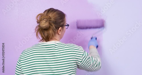 Young tired woman listlessly paints wall with paint roller in lilac color. Color therapy influence of color on human condition Lady indifferently paints the wall purple, she is tired, depressed. photo