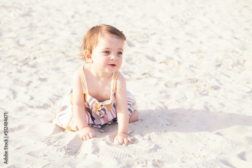happy baby girl sitting on the beach on sand wearing striped summer dress on sunny day