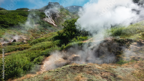 Steam and smoke rise from the fumarole on the hillside. Sulphurous deposits on the soil. Green vegetation on the mountains. Blue sky. Kamchatka. Valley of Geysers