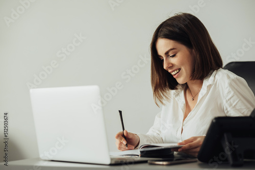 Cheerful Young Woman Working at the Office by Laptop, Female Accountant Writing Notebook at Workplace