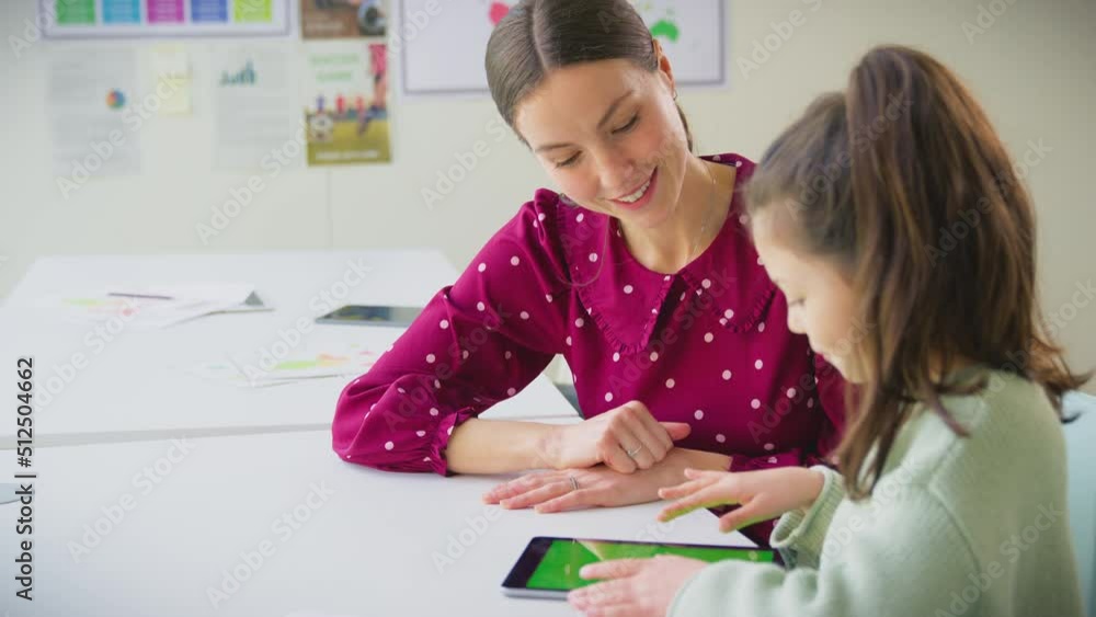 Female teacher sitting at desk with female elementary pupil in classroom using digital tablet in school classroom together- shot in slow motion Female teacher sitting at desk with female elementary pupil in classroom using digital tablet in school classroom together- shot in slow motion