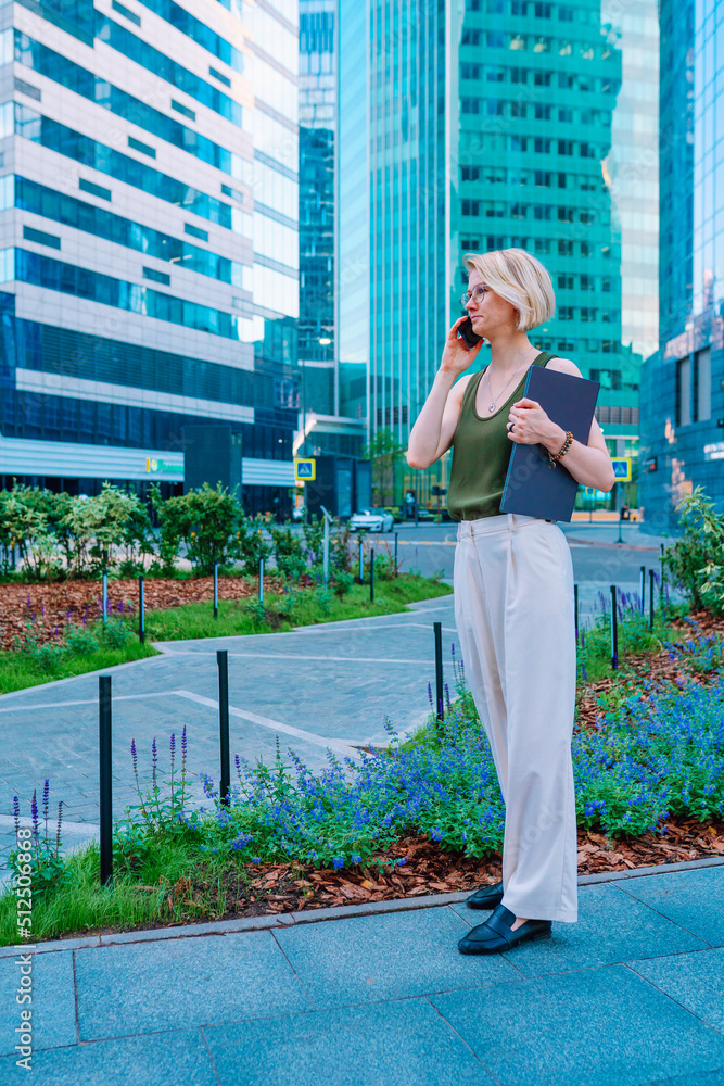 Side view of middle-aged blond woman businesswoman wearing green top, white trousers, standing on street in city centre near flowered beds, skyscrapers, holding blue laptop, talking on smartphone.