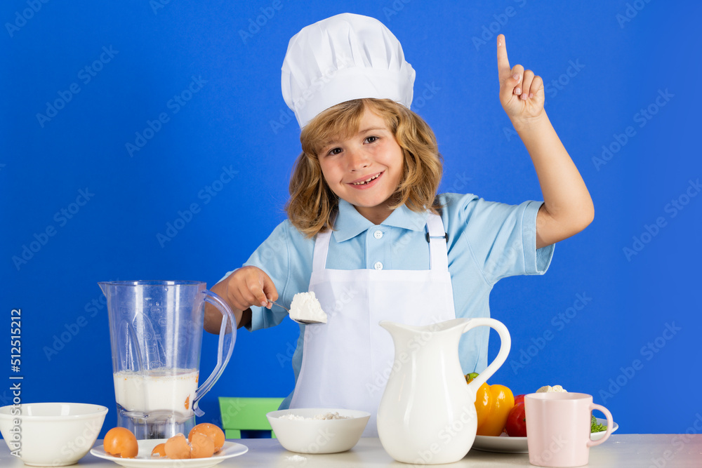 Child chef cook prepares food with flour in isolated blue studio background. Kids cooking. Teen boy with apron and chef hat preparing a healthy vegetables meal in the kitchen. Child chef cook prepares food with flour in isolated blue studio background. Kids cooking. Teen boy with apron and chef hat preparing a healthy vegetables meal in the kitchen.