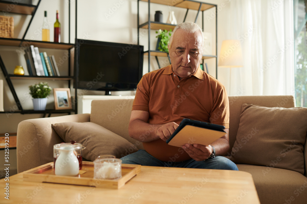 Senior man drinking tea and reading news article on tablet computer at home Senior man drinking tea and reading news article on tablet computer at home