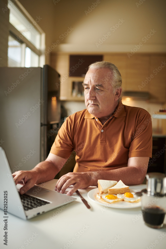 Elderly man checking his social media account on laptop when eating breakfast at home Elderly man checking his social media account on laptop when eating breakfast at home