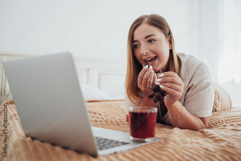 Beautiful Caucasian Woman Laying on the Bed in Hotel Room and Eating Grapes While Watching TV Series Through Laptop Beautiful Caucasian Woman Laying on the Bed in Hotel Room and Eating Grapes While Watching TV Series Through Laptop