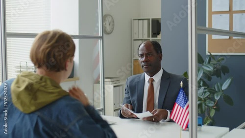 African American consul checking documents and speaking with Caucasian visa applicant during interview in US embassy
