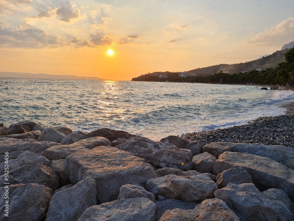 Scenery with sunset on the beach. Sunset over the sea. Scenic landscape with surf on waves in the evening. Beautiful sun rays at sunset. Time lapse twilight. Panoramic view of pebble beach and stones.