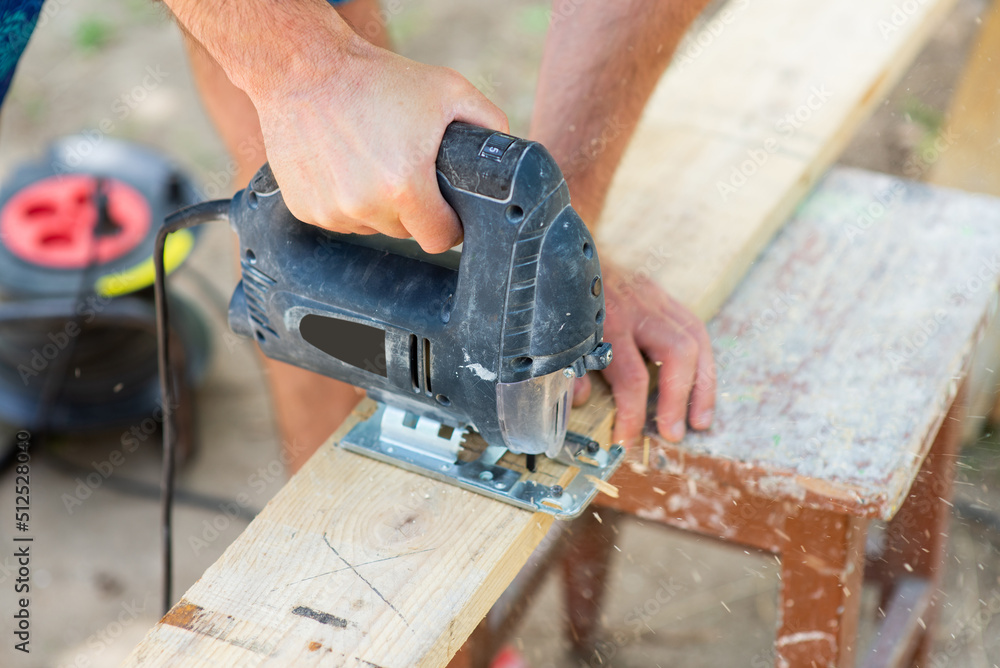 A male carpenter cuts a wooden plank with a jigsaw, close-up