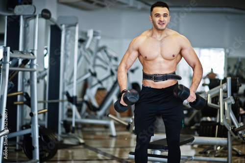 Muscular arab man training in with dumbbells modern gym. Fitness arabian men with naked torso doing workout .