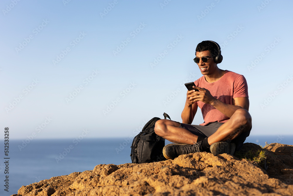 Young man sitting on the stone listening the music. Tourist man on the top of the mountain