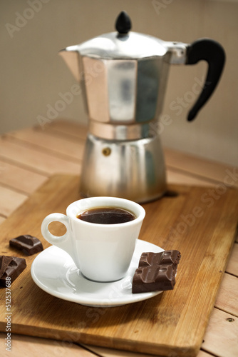 White cup of espresso coffee with piece of chocolate on plate and vintage geyser coffee maker on background