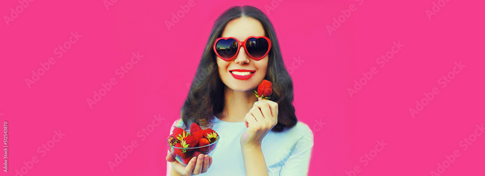 Portrait close up of happy smiling woman with handful of fresh strawberries wearing red heart shaped sunglasses on pink background Portrait close up of happy smiling woman with handful of fresh strawberries wearing red heart shaped sunglasses on pink background