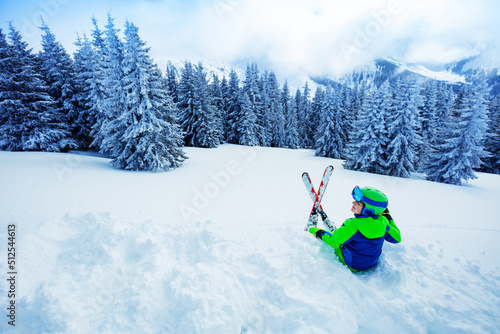 Boy sit in snow with ski over fir forest after heavy snowfall