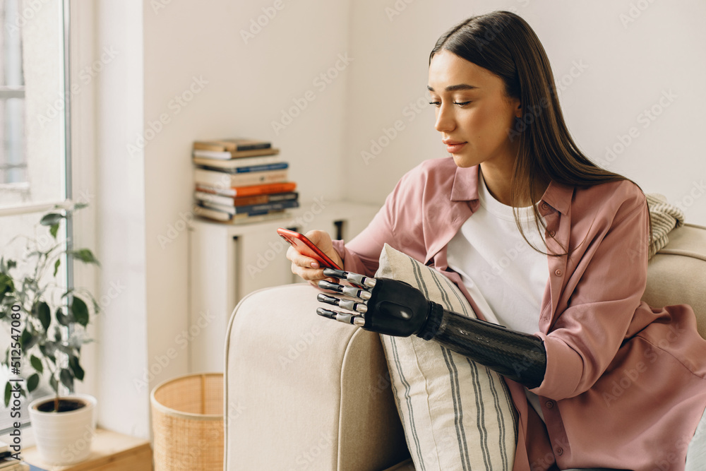 Person with special needs. Attractive young Caucasian woman sitting on couch using red smartphone, holding it in bionic black 3d-printed hand, wearing stylish pink silk shirt, having lovely face Person with special needs. Attractive young Caucasian woman sitting on couch using red smartphone, holding it in bionic black 3d-printed hand, wearing stylish pink silk shirt, having lovely face