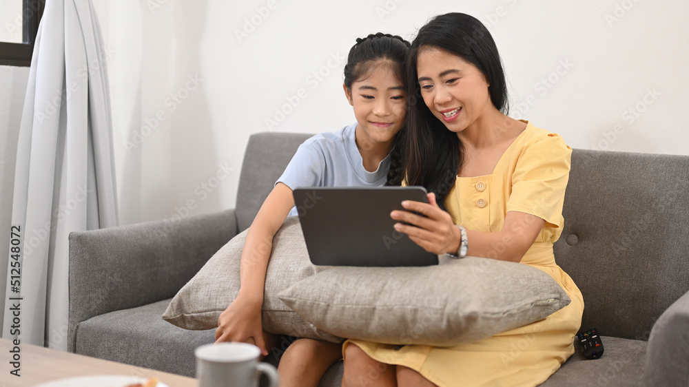 Smiling asian mom and daughter having fun browsing internet on digital tablet, enjoying spending free time together Smiling asian mom and daughter having fun browsing internet on digital tablet, enjoying spending free time together