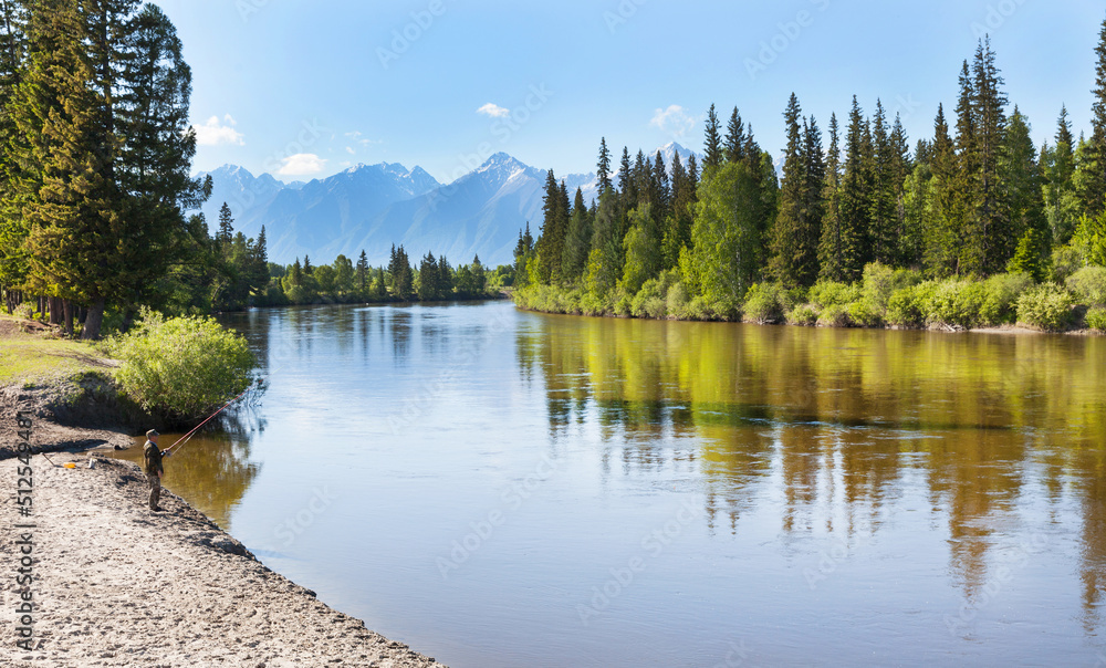 Beautiful summer landscape with a green forest on the banks of the Irkut River and the mountain peaks of the Eastern Sayan Mountains on sunny June day. Village fisherman with fishing rod on the shore Beautiful summer landscape with a green forest on the banks of the Irkut River and the mountain peaks of the Eastern Sayan Mountains on sunny June day. Village fisherman with fishing rod on the shore