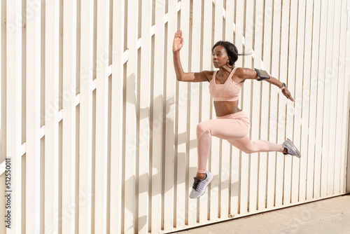 Young African American sporty woman jumping up and running against light background. Female model in sportswear exercising outdoors photo