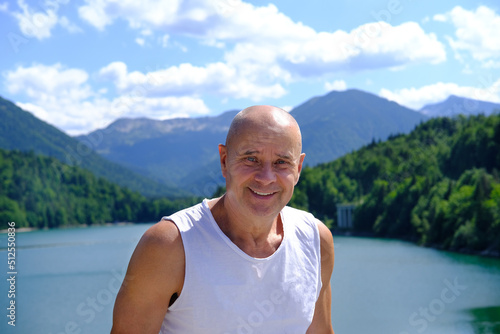 mature man of 60 years old stands, smiles against backdrop of lake Achensee in Austria, green mountains rises above calm expanse of water, concept of vacation by reservoir, resort place tyrol