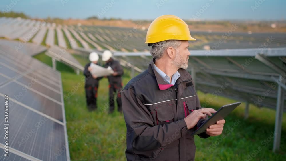 Senior Caucasian engineer in protective helmet and uniform stands in field with solar panels looking for information in tablet on background of colleague with paper diagram. Senior Caucasian engineer in protective helmet and uniform stands in field with solar panels looking for information in tablet on background of colleague with paper diagram.