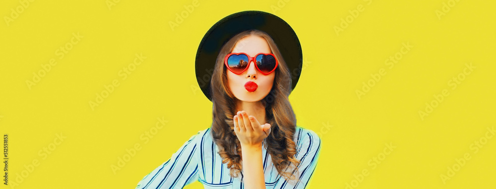 Portrait of beautiful young woman blowing her lips sending air kiss wearing red heart shaped sunglasses, black round hat, striped t-shirt on yellow background, blank copy space for advertising text