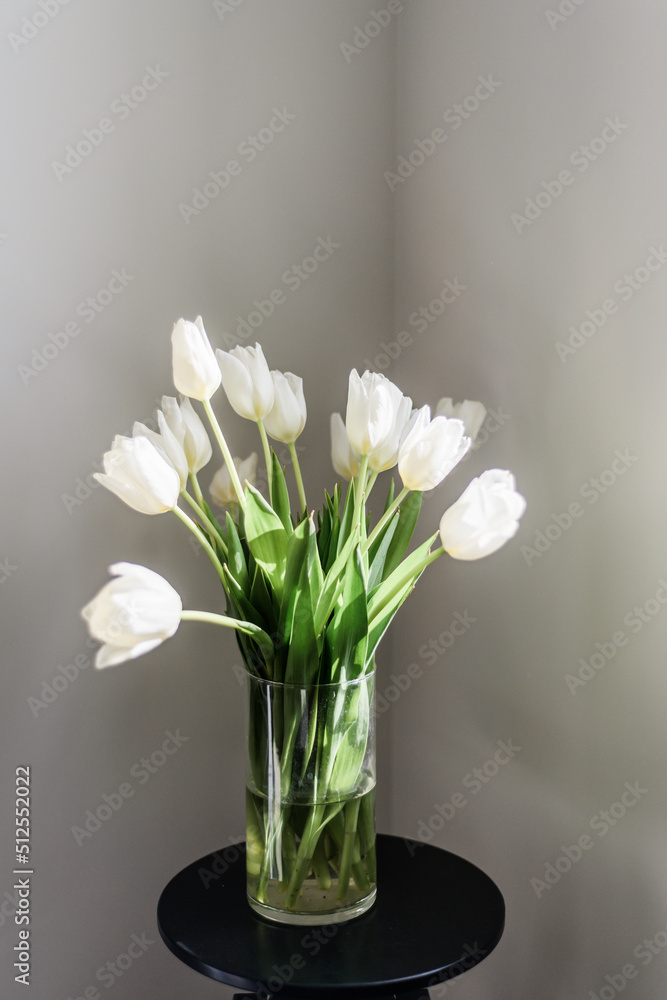 A bouquet of beautiful white tulips in a clear vase stands on a blue table. A light wall behind and a place for the text. Home interior