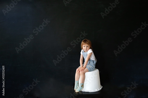 Sad European girl sitting on white bucket on black background, copy space