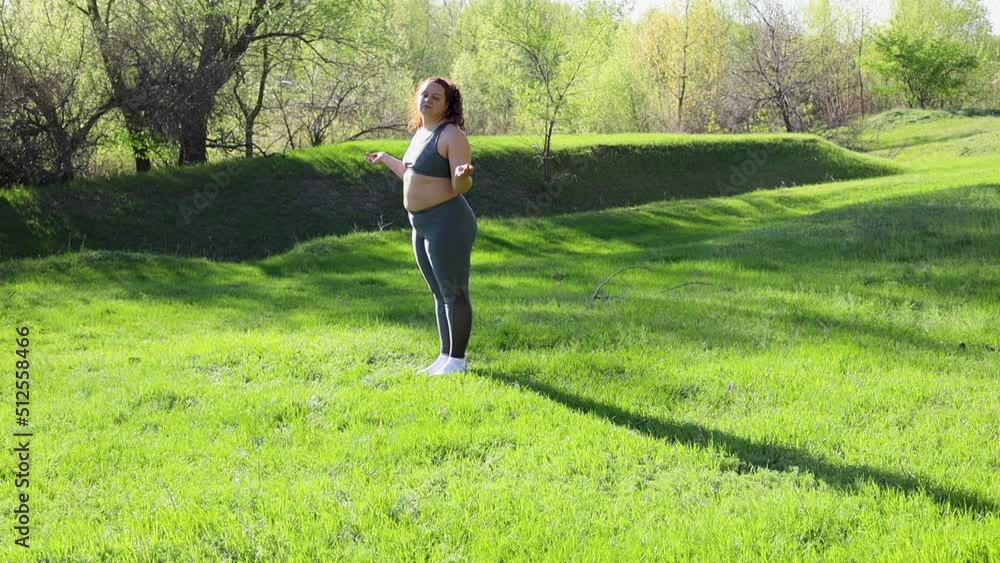 Young overweight woman in fitness suit turning head standing on green lawn in park on sunny summer day, slow motion. Healthy lifestyle, exercise and fitness outdoors. Sports activity concept. Young overweight woman in fitness suit turning head standing on green lawn in park on sunny summer day, slow motion. Healthy lifestyle, exercise and fitness outdoors. Sports activity concept.