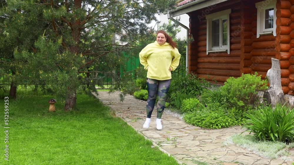 Red-haired curly plus size fat female with excess weight in yellow hoodie do cardio exercises, fitness, bouncing and jumping from side to side in green backyard. Nature, meadow and house on background Red-haired curly plus size fat female with excess weight in yellow hoodie do cardio exercises, fitness, bouncing and jumping from side to side in green backyard. Nature, meadow and house on background