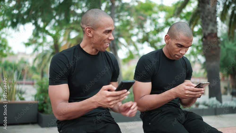 Two laughing bald African brothers looking at phone while sitting on the bench outdoors Two laughing bald African brothers looking at phone while sitting on the bench outdoors