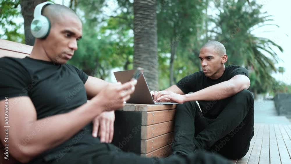 Concentrated bald African brothers typing on phone and working on laptop while sitting on the bench outdoors Concentrated bald African brothers typing on phone and working on laptop while sitting on the bench outdoors