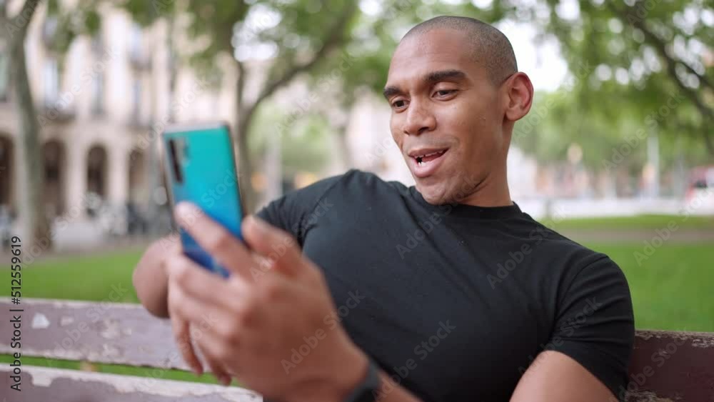 Laughing bald African man talking by video call on phone while sitting on the bench outdoors Laughing bald African man talking by video call on phone while sitting on the bench outdoors