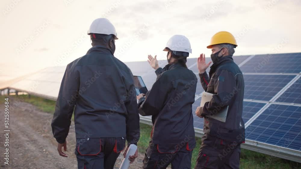 Three engineers in protective face masks, hard hats and uniforms walk among rows of solar panels and communicate. View from back. Three engineers in protective face masks, hard hats and uniforms walk among rows of solar panels and communicate. View from back.